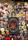 Kulture Vulture logo black coozie featuring the vulture logo in red and the words 'Kulture Vulture New Orleans' along with the website URL in white text, photographed on a bottle on top of a bunch of band button pins.