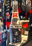 Rush Neil Peart RIP coozie featuring a black and white image of him on the drums with his name across the top and the dates "1952-2020" across the bottom in black and white text, photographed on a beer bottle.