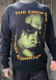 A person wearing a black long-sleeve graphic t-shirt featuring a large green-toned face print of Brandon from The Crow movie and orange text reading 'THE CROW' and 'NOTHING IS TRIVIAL'.