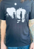 The Cure Exorcist black t-shirt featuring a silhouette of Robert Smith in the style of the Exorcist movie imagery, photographed on a model in front of a metal gate background.