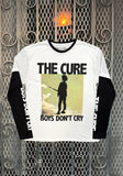 A white and black long-sleeve graphic t-shirt featuring the 'The Cure - Boys Don't Cry' album artwork, displayed against a metal gate background.