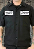 Black button down work shirt featuring an embroidered patch with a white background and black words 'Customer Service' on the right breast and an embroidered patch with a white background and black words 'I Don't Really Give a Shit' on the left breast, on a model against a metal gate background.
