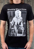 Dolly Parton Guitar black t-shirt featuring a black and white framed image of her posing with her guitar and white text 'Dolly Parton' above the image, photographed on a model in front of a metal gate.
