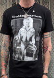 Dolly Parton Guitar black t-shirt featuring a black and white framed image of her posing with her guitar and white text 'Dolly Parton' above the image, photographed on a model in front of a metal gate.