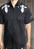 Front of black button down work shirt featuring black and white images of Elvira looking surprised on each upper shoulder and the word 'Elvira' in white script on the bottom of the left sleeve, on a model against a metal gate background.