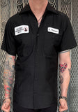 Black button down work shirt featuring an embroidered patch with a white background and black script 'F. Krueger' on the left breast and an embroidered patch with a white background and black text 'Elm Street Neighborhood Watch' on the right breast, on a model against a metal gate background.