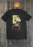 Iggy Pop I Wanna Be Your Dog black t-shirt featuring large yellow text 'Now I Wanna Be Your Dog' above an image of Iggy crawling on all fours with his tongue sticking out, displayed on a metal gate background.