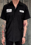 Black button down work shirt featuring an embroidered patch with a white background and black script 'Jason' on the left breast and an embroidered patch with a white background and text stylized in wood planks 'Camp Crystal Lake' on the right breast, on a model against a metal gate background.