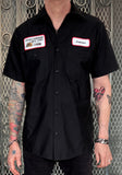 Black button down work shirt featuring an embroidered patch with a white background, red trim and black script 'Leatherface' on the left breast and an embroidered patch with a white background, red trim and text 'Texas Chainsaw Mfg. Corp.' along with an image of a chainsaw on the right breast, on a model against a metal gate background.