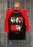 The Lost Boys black raglan t-shirt with red 3/4 sleeves featuring the movie imagery along with red text that reads "Sleep all day Party all night Never grow old Never die The Lost Boys", photographed flat on a metal gate background.