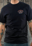 Front of Lucky 13 Frankie's Garage black t-shirt featuring the logo over the left chest, on a model against a metal gate background.