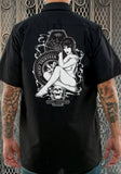 Back of Lucky 13 Miss Trust black work shirt featuring an image of a lady, a spiderweb on a coffin, a tire, and a skull, photographed on a model in front of a metal gate.