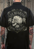 Back of black button down work shirt featuring stylized text 'Dia De Los Muertos Since 1991 Lucky Thirteen' accented with four winged skull and crossbones around a large centered side facing, tattooed skull illustration with roses around it all in shades of off white with bronze accents, on a model against a metal gate background.