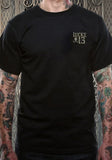 Front of Lucky 13 Muertos black t-shirt featuring a Lucky 13 logo with skull and crossbones on the left chest, on a model against a metal gate background.