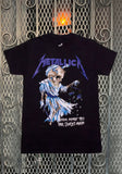 Front of Metallica Harvester of Sorrow black t-shirt featuring an illustration of the Harvester along with the band logo across the top in purple and the text 'their money tips her scales again' in white on the lower left, displayed on a metal gate background.