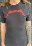 Metallica Kill 'Em All Classic Logo black t-shirt featuring the classic logo in red across the chest, on a model against a metal gate background.