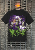 Munsters Spooky Family black t-shirt featuring the word 'Musters' in large green dripping text along with a colorful image of the family, displayed on a metal gate background.