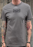 Nine Inch Nails grey t-shirt featuring a puffy NIN logo in the upper middle of the chest, photographed on a model against a metal gate background.