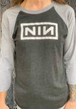 Nine Inch Nails heathered black raglan t-shirt with grey 3/4 sleeves featuring the NIN logo, photographed on a model in front of a metal gate background.