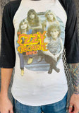 Ozzy Osbourne band white raglan t-shirt with black 3/4 sleeves featuring yellow and orange words 'the Ozzy Osbourne band' and an image of the 4 of them sitting on steps, on a model in front of a metal gate background.