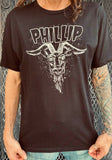 Black Phillip The Witch black t-shirt featuring a black and white illustration of Phillip with the word "Phillip" in large black letters outlined in white at the top, on a model in front of a metal gate background.