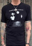 A person wearing a black short-sleeve graphic T-shirt with white text reading 'THE SISTERS OF MERCY - FLOODLAND' and a large black-and-white band photo print on the front.