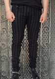 Male model wearing skinny leg fitted, 5 pocket jeans style dark charcoal pants with distressed black vertical stripes, against a metal gate background.