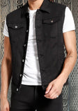 Front of black denim vest featuring metal buttons down the front and on the chest pockets, worn open on a model against a metal gate background.
