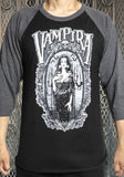 Vampira black raglan t-shirt with dark charcoal heather grey 3/4 sleeves featuring a black and white image of her inside an elaborate frame, on a model against a metal gate background.