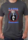 Frank Zappa for President grey t-shirt featuring an illustration of his head wearing a patriotic top hat circled in red and blue stars with the text 'Zappa for President' around the image, on a model against a metal gate background.