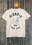 Blondie NYC white t-shirt featuring a distressed illustration of a woman in a bikini lounging and smoking with the words "Blondie 1974 New York" around her, photographed flat on a metal gate background.