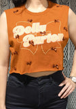 A woman wearing a sleeveless cropped copper top with a large retro-style 'Dolly Parton' graphic in cream and white, decorated with small brown bow embellishments, paired with black jeans.