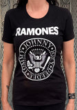 Ramones black t-shirt featuring the classic logo with a graphic image of an eagle in the middle and "Johnny Joey DeeDee Tommy" in a circle around it and the Ramones logo across the top, photographed on a model in front of a metal gate background.