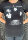 A person wearing a black short-sleeve graphic T-shirt with white text reading 'THE SISTERS OF MERCY : FLOODLAND' and a large white moon-and-waterfront print on the front, photographed against a metal mesh gate.