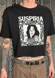 A person wearing a black cropped graphic T-shirt with the word 'SUSPIRIA' in bold white letters above a black-and-white portrait print, standing in front of a metal gate.