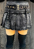 A black distressed denim pleated mini skirt with a wide studded belt, shown on a model against a metal mesh background.