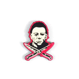 Close-up of Halloween Movie Franchise enamel pin featuring head shot of Michael Myers with crossed knives dripping blood beneath him, outlined in a pinkish red color, photographed on a white background.