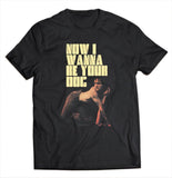 Close-up of Iggy Pop I Wanna Be Your Dog black t-shirt featuring large yellow text 'Now I Wanna Be Your Dog' above an image of Iggy crawling on all fours with his tongue sticking out, displayed on a white background.